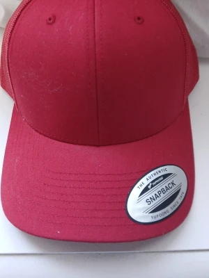 Röd snapbackkeps från Yupoong - Snygg röd snapbackkeps från Yupoong, modell The Classics. Kepsen har böjd skärm, mesh på sidorna och justerbar snapback baktill. Perfekt för en streetwear-look eller att lyfta din outfit med en färgklick.