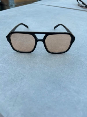Svarta retro solglasögon med brun ton - Coola solglasögon med svart, kantig båge och bruna glas. Modellen har en retroinspirerad design med markerad näsbrygga och stora rutor. Perfekta för dig som vill sticka ut med en unik accessoar i sommar. 🕶️☀️⛱️