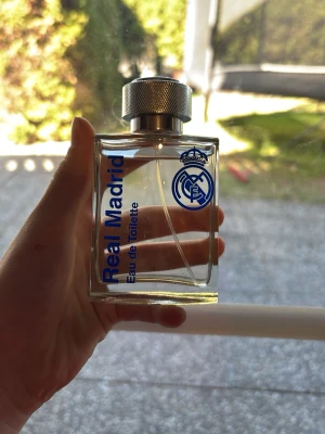 Real Madrid Eau de Toilette - Fräsch parfym i en genomskinlig, fyrkantig glasflaska med silverfärgad metallkork. Flaskan har blå text och Real Madrids logga tryckt på framsidan. Perfekt för dig som gillar sportiga dofter och vill visa ditt stöd för Real Madrid. Ca 99ml av 100ml kvar