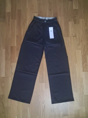 Svart Suit Up mjukisbyxor från Alo Yoga - Svart mjukisbyxor från Alo med breda ben och hög midja. Byxorna har elastisk midja och diskret Alo-logga på ena benet. Tillverkade i ett mjukt och följsamt material som känns skönt mot huden. Perfekta för en avslappnad och trendig look.