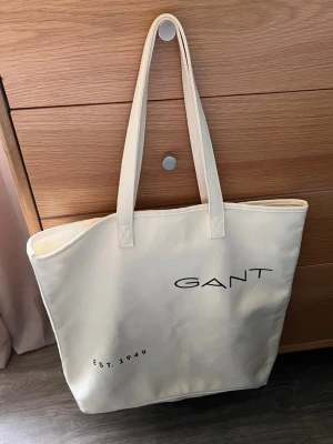 GANT shoppingväska - Stilren shoppingväska i beige canvas från GANT med svarta tryckta detaljer. Väskan har två långa handtag och texten 'GANT' samt 'EST. 1949' på framsidan. Perfekt storlek för att bära med sig allt man behöver under dagen. Storlek 46x46 cm. Använd fåtal gånger.