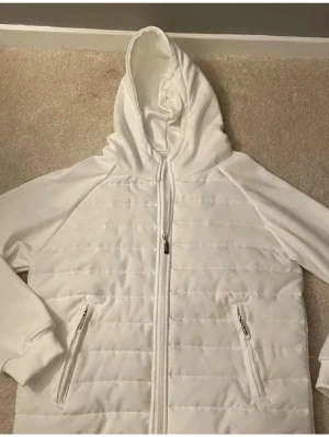 Vit hoodie med quiltad front - Säljer en vit hoodie med quiltad front och dragkedja. Hoodien har två fickor med dragkedja och en stor huva. Ärmarna och ryggen är i mjukt tyg medan framsidan är quiltad för extra stil. Perfekt för dig som gillar en clean look.