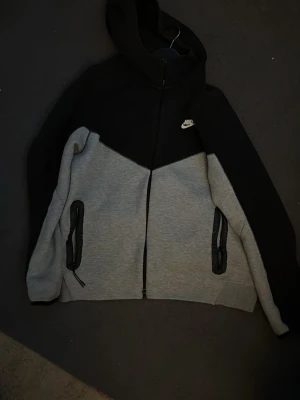 Nike tvåfärgad hoodie - Snygg hoodie från Nike i svart och grått med dragkedja framtill och två fickor med svarta detaljer. Klassisk Nike-logga på bröstet och huva för extra stilpoäng. Perfekt för en sportig och avslappnad look.