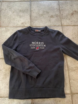 Svart tröja från Morris - Svart långärmad tröja från Morris Stockholm med broderad logga och text på bröstet. Tröjan har rund halsringning och ribbade muddar vid ärmslut och nederkant. Perfekt för en stilren och klassisk look.