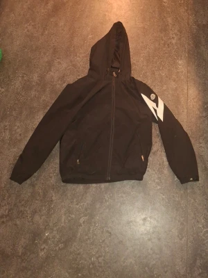 Svart moncler vindjacka med huva och tryck - Svart vindjacka med huva och dragkedja framtill. Jackan har två fickor med dragkedja och ett vitt grafiskt tryck på ena ärmen. Tillverkad i lätt och slitstarkt material, perfekt för blåsiga dagar. Trycket hänger lite löst men kan fixas med strykjärn.