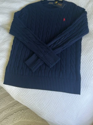 Mörkblå kabelstickad tröja Polo Ralph Lauren - Kabelstickad mörkblå tröja från Polo Ralph Lauren med rund halsringning och röd broderad logga på bröstet. Tröjan har ribbade muddar och nederkant, perfekt för lager-på-lager och snygg till jeans.