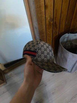 Gucci beige keps - Säljer en beige keps från Gucci med klassiskt GG-monogram över hela kepsen och en röd-blå rand på sidan. Kepsen har böjd skärm och justerbar baksida. Materialet är canvas med detaljer i svart. Perfekt för dig som gillar exklusiva accessoarer.