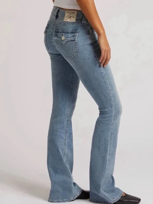 Ljusblå bootcut jeans True Religion - Säljer ett par ljusblå bootcut jeans från True Religion med klassiska sömmar och snygga fickdetaljer bak. Jeansen har låg midja och en ikonisk patch på linningen. Perfekta för dig som gillar en avslappnad men trendig look. De är lite sönder i nederkant på grund av att man trampat på dem.
