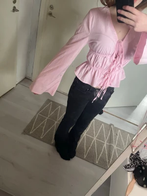 Ljusrosa blus med knytning - Supersöt ljusrosa blus med v-ringning och knytning framtill. Blusen har långa, vida ärmar och markerad midja med volangkant. Perfekt för en trendig och feminin look. Materialet ser ut att vara lätt och luftigt, vilket ger en skön känsla.