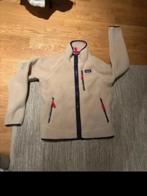 Beige fleecejacka från Patagonia - Mysig beige fleecejacka från Patagonia med marinblå dragkedjor och detaljer i rött. Jackan har hög krage, tre fickor med dragkedja och en klassisk logga på bröstet. Perfekt för lager-på-lager och chill höstkvällar.