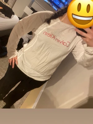 Vit långärmad topp från Calvin Klein - Vit långärmad topp från Calvin Klein med röd logga framtill. Klassisk rund halsringning och rak passform. Perfekt för en avslappnad och stilren look.