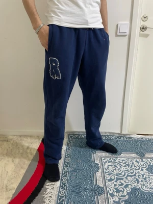 Mörkblå Risky-byxor  - Säljer ett par mörkblå mjukisbyxor med broderad björnpatch på vänster ben. Byxorna har dragsko i midjan och muddar vid bensluten. Perfekta för chill dagar hemma eller när du vill ha en avslappnad stil.