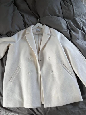 Oversized vit ullkappa - Stilren oversized kappa i vit ullmix med dubbelknäppning och breda slag. Jackan har en rak passform och släta knappar framtill. Perfekt för dig som gillar minimalistisk och trendig stil.