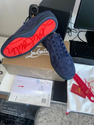 Mörkblå Christian Louboutin sneakers - Snygga mörkblå sneakers från Christian Louboutin i mocka med klassisk röd sula och diskret logga på sidan. Skorna har lågt skaft, svarta skosnören och stilren design som sticker ut med den ikoniska röda undersidan.