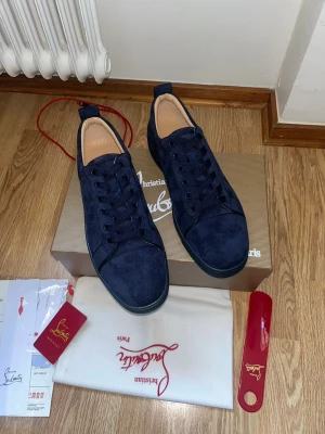 Mörkblå sneakers från Louboutin - Snygga mörkblå sneakers från Christian Louboutin i mocka med klassisk röd sula och svarta snören. Låg modell med rund tå och diskreta sömdetaljer. Perfekta för dig som vill sticka ut med exklusiv stil.