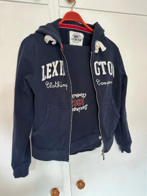 Mörkblå hoodie från Lexington - Mörkblå hoodie med dragkedja från Lexington. Stor broderad logga framtill, vita snören i huvan och kängurufickor. Mjuk bomullskvalitet och klassisk college-stil. Zip-Hoodie i Storlek S och byxorna i M. Älskar detta set, men tyvärr för stort för mig 🌸