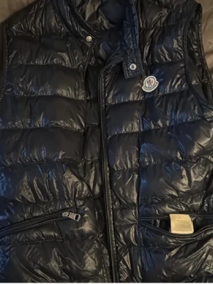 Mörkblå dunväst från Moncler - Snygg mörkblå dunväst från Moncler med glansig finish och klassisk logga på bröstet. Västen har ståkrage, dragkedja framtill och två fickor med dragkedja. Perfekt för lager-på-lager och streetstyle.