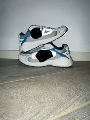 Vita och blå sneakers med meshdetaljer - Snygga sneakers i vitt med blå och grå detaljer. Skorna har en sportig design med meshpaneler för extra andningsförmåga och vita snören. Sulan är vit med svarta partier undertill och skon har en låg profil som passar till många stilar.