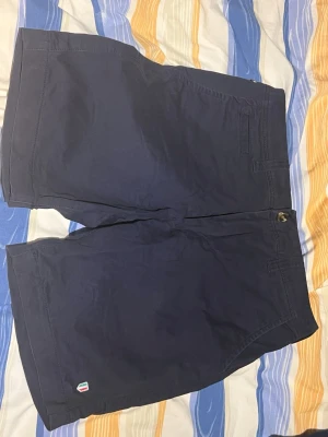 Mörkblå shorts från Strömberg - Mörkblå shorts från Strömberg med klassisk design och diskret logga vid benet. Tillverkade i ett mjukt och bekvämt bomullsmaterial, perfekta för varma dagar. Stängs med knapp och dragkedja framtill och har fickor på sidorna.