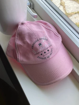Ljusrosa keps med broderad text - Säljer en ljusrosa keps från Genuine Design med broderad text och små stjärnor framtill. Kepsen har böjd skärm och justerbart spänne baktill i metall. Tillverkad i bomullstyg och passar perfekt för en avslappnad stil.