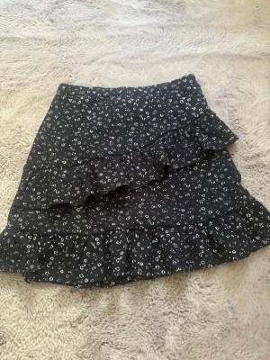 Svart blommig volangkjol - Snygg svart kortkjol med små vita blommor och volangdetaljer. Kjolen har en feminin och trendig look, perfekt för dig som gillar en söt och lekfull stil. Materialet känns lätt och bekvämt.