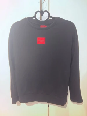 Svart sweatshirt från Hugo Boss - Svart sweatshirt med rund hals från Hugo Boss. Tröjan har en röd patch med Hugo-loggan på bröstet och är tillverkad i mjukt material. Långärmad och med ribbade muddar vid ärmslut och nederkant. Perfekt för en avslappnad och stilren look.