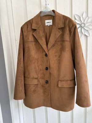 Brun kavaj i mockaimitation från ONLY - Snygg brun kavaj i mockaimitation från ONLY med klassisk krage, tre svarta knappar och två fickor framtill. Rak passform och helfodrad för en stilren look. Perfekt för dig som vill ha en trendig och tidlös kavaj i garderoben.