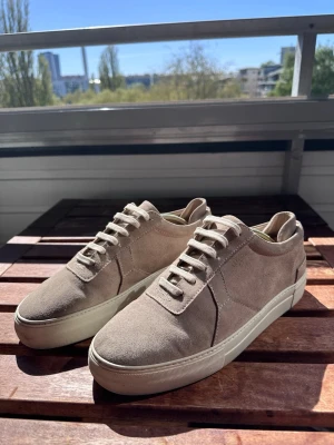 Beigea sneakers från Axel Arigato - Stilrena sneakers från Axel Arigato i beige mocka med matchande beige skosnören och vit gummisula. Klassisk låg modell med diskret design och dragflik bak. Perfekta för en clean och avslappnad look.