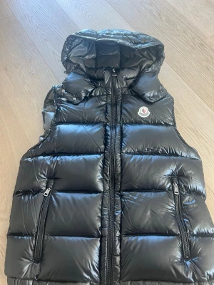 Svart dunväst från Moncler med huva - Snygg svart dunväst från Moncler med glansig finish, avtagbar huva och två dragkedjefickor framtill. Västen har Moncler-logga på bröstet och breda, vadderade paneler för extra värme. Perfekt för lager-på-lager och streetstyle.