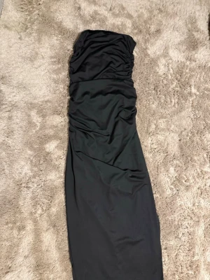 Svart axelbandslös långklänning - Snygg svart axelbandslös långklänning med figurnära passform och lätt rynkade detaljer upptill plus öppning i ena benet. Klänningen har en stilren och elegant look. Materialet känns mjukt och följsamt mot kroppen. Obs: kommer från Shein. Säljs pga: använder aldrig 