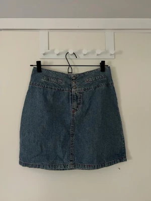 Blå jeanskjol med knappar - Snygg blå jeanskjol i klassisk modell med tre knappar framtill och fickdetaljer. Ganska högmidjad men beror på hur man vill att den ska sitta