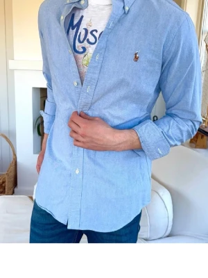Ralph Lauren skjorta - Klassisk ljusblå skjorta från Ralph Lauren med button-down krage och broderad logga på bröstet. Skjortan är långärmad och har vita knappar framtill. Perfekt för en stilren och avslappnad look.
