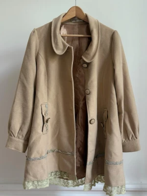 jfashion kappa - Snygg beige kappa från det kända japanska märket axes femme, rundad krage och dekorativa knappar. Fina spetsdetaljer och broderiband längs nederkant och rygg. Puffiga ärmar med knappslå och rosettdekor vid fickorna. jättebra skick men endast lite skrynklig som kan lätt fixas. japansk storlek M men sitter som EU XS-S/ 36 paketpris om du köper mer