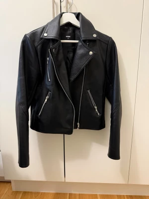 Svart skinnjacka från Bik Bok - Snygg svart skinnjacka från Bik Bok med klassisk bikerstil, silvriga dragkedjor och tryckknappar. Jackan har bred krage och två snedställda fickor framtill. Perfekt för en cool och trendig look.