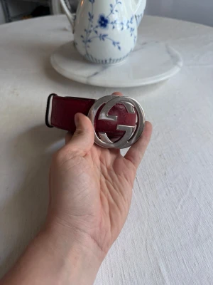 Rött skärp från Gucci - Snyggt rött skärp från Gucci i skinn med präglad logga längs hela bandet och en stor, rund silverspänne med GG-logga. Perfekt accessoar för att lyfta din outfit med en lyxig touch. Passar midja 30-33. Kan göras mindre med mer hål.