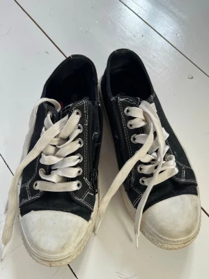 Svarta låga sneakers från DinSko - Klassiska svarta sneakers från DinSko med vita gummisulor och tå, vita snören och kontrasterande vita sömmar. Skorna har en låg profil och är tillverkade i canvasmaterial, perfekta för en avslappnad streetstil.