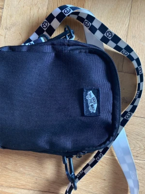 Svart axelväska från Vans - Cool svart axelväska från Vans med vit och svart schackrutigt axelband med peace-symboler. Väskan har dragkedja och är tillverkad i slitstarkt tygmaterial. Perfekt för att bära det viktigaste och ger en streetstyle-vibe.
