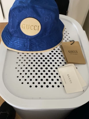 Blå bucket hat från Gucci - Cool blå bucket hat från Gucci med diskret GG-mönster i tyget och ett stort beige läderpatch med guldfärgad logotyp framtill. Hatten har beige kant och är tillverkad i ett slitstarkt material som passar perfekt för att lyfta din stil.