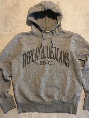 Grå hoodie Replay Blue Jeans - Snygg grå hoodie från Replay Blue Jeans med stor logga framtill. Klassisk modell med huva och dragsko, ribbade muddar vid ärmar och midja. Tillverkad i mjukt sweatshirtmaterial som är skönt att ha på sig. Perfekt för en avslappnad och trendig look.