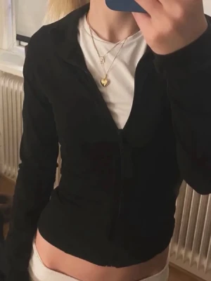 Svart tröja från Gina  - Snygg svart kofta med v-ringning och långa ärmar. Perfekt att bära över en t-shirt för en enkel men stilren look. Koftan har en figurnära passform och knäppning framtill.