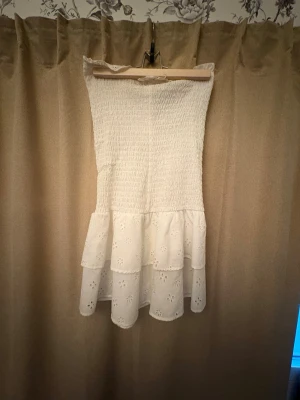 Vit axelbandslös sommarklänning - Söt vit axelbandslös klänning med smockad överdel och volangig kjol med broderade blommor. Perfekt för varma dagar och har en luftig känsla. Klänningen är kort och har en romantisk stil. Inga skavanker!!