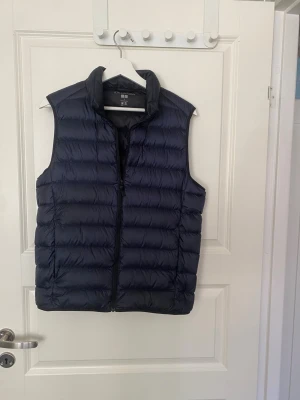 Mörkblå dunväst från Uniqlo - Snygg mörkblå dunväst från Uniqlo Ultra Light Down-serien. Västen är lätt, har vattenavvisande material och dragkedja framtill. Perfekt för lager-på-lager och enkel att bära med sig. Klassisk quiltad design och hög krage för extra värme.