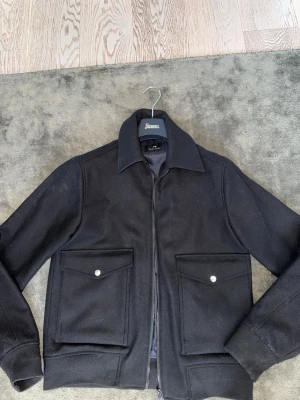 Svart jacka från Paul Smith - Stilren svart jacka från Paul Smith med klassisk krage, stora fickor framtill med knappar och dragkedja. Jackan har en rak passform och är fodrad med glansigt tyg. Perfekt för dig som gillar minimalistisk och tidlös stil.