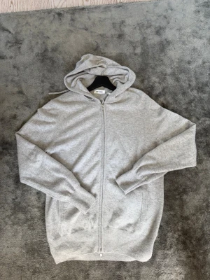 Ljusgrå hoodie från Soft Goat - Mysig ljusgrå hoodie från Soft Goat i mjuk kashmirblandning. Dragkedja framtill, två fickor och ribbade muddar. Perfekt för en chill och stilren look. Klassisk huva med dragsko och enkel design som passar till det mesta.