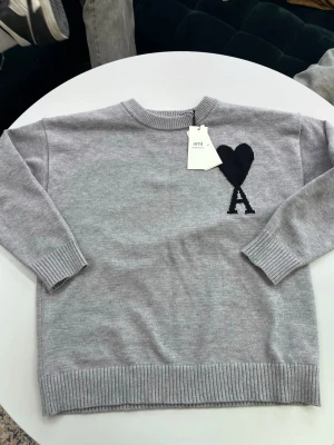 Grå stickad tröja från Ami Paris - Snygg grå stickad tröja från Ami med svart hjärta och bokstaven A broderat på bröstet. Tröjan har rund halsringning, ribbade muddar och nederkant. Perfekt för dig som gillar stilrena och trendiga plagg.