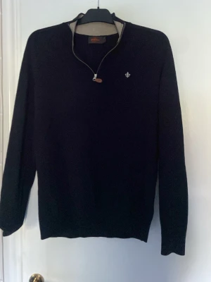 Mörkblå stickad tröja med halvzip - Mörkblå stickad tröja från Morris med halvzip och en liten logotyp broderad på bröstet. Tröjan har lång ärm, ribbade muddar och en klassisk passform. Perfekt för lager-på-lager och snygg till jeans. Materialet är mjukt och behagligt. storlken är m men passar även s 