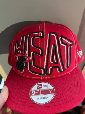 Röd Miami Heat keps från New Era - Snygg röd snapback-keps från New Era med stor broderad HEAT-text och Miami Heat-logga framtill. Kepsen har platt skärm, justerbar snapback och broderade detaljer. Perfekt för dig som gillar basket och streetwear.