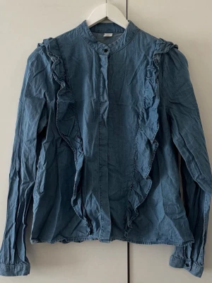 Blå volangblus med långa ärmar - Supersnyggw blå blus med volangdetaljer framtill och längs axlarna. Blusen har en rak passform, ståkrage och knäppning framtill. Tillverkad i ett mjukt, jeansliknande material som ger en avslappnad men trendig look.