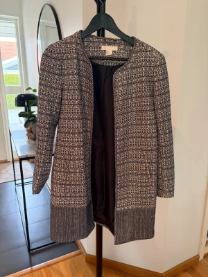 Mönstrad svartvit kappa från H&M - Stilren svartvit kappa från H&M med grafiskt mönster och rund halsringning. Kappan är öppen framtill, har långa ärmar och bredare enfärgad kant nedtill. Perfekt för dig som gillar en klassisk men trendig look.