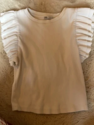 Vit volangtopp från Zara - Supersöt vit topp från Zara med volangdetaljer på axlarna. Toppen har rund halsringning och är tillverkad i mjuk bomull. Perfekt för dig som vill ha en enkel men ändå trendig look. Typ en magtröja